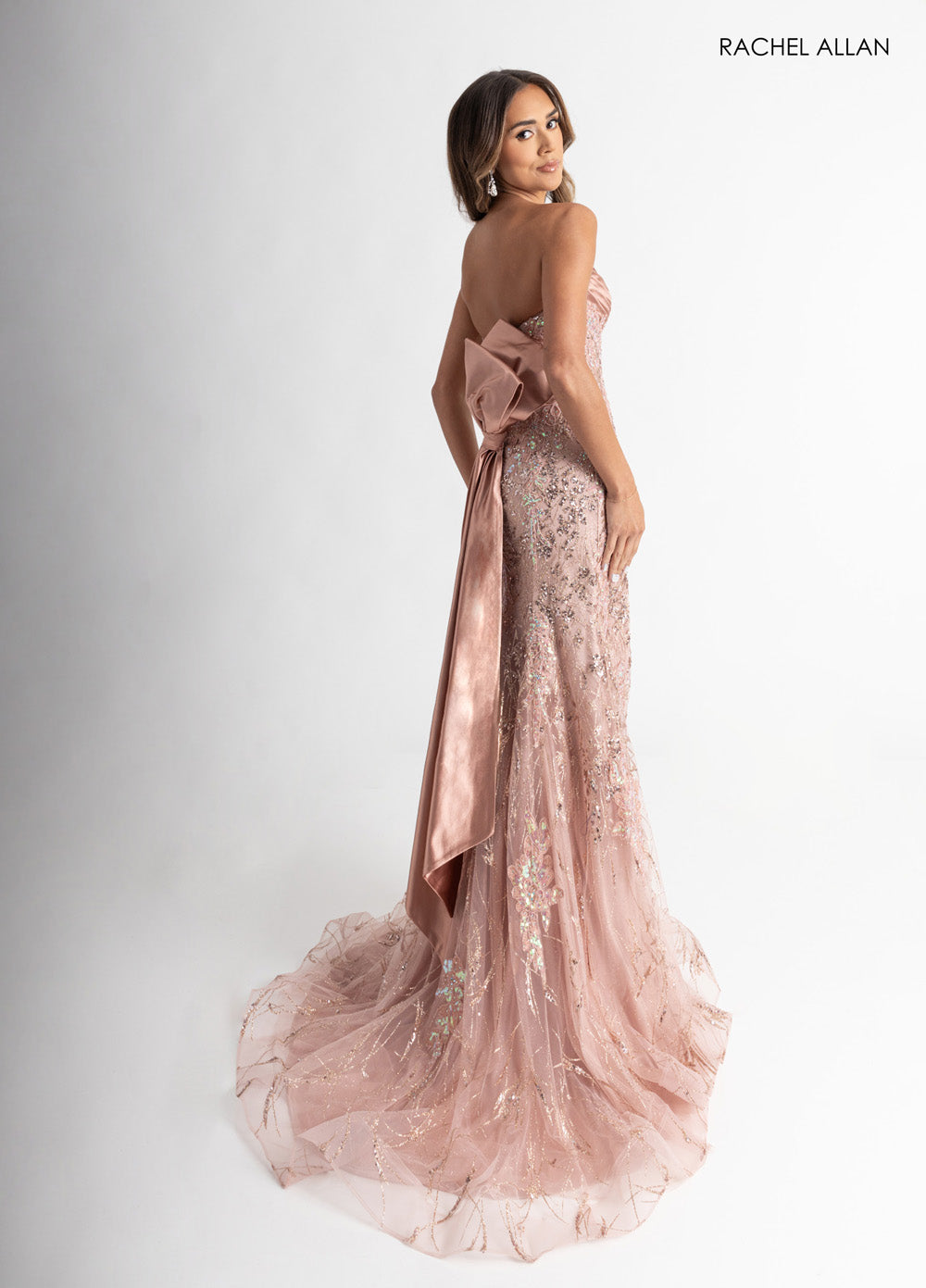 Rachel Allan 71008 Rose gold formal gown with crystal beading and a dramatic bow, back view.