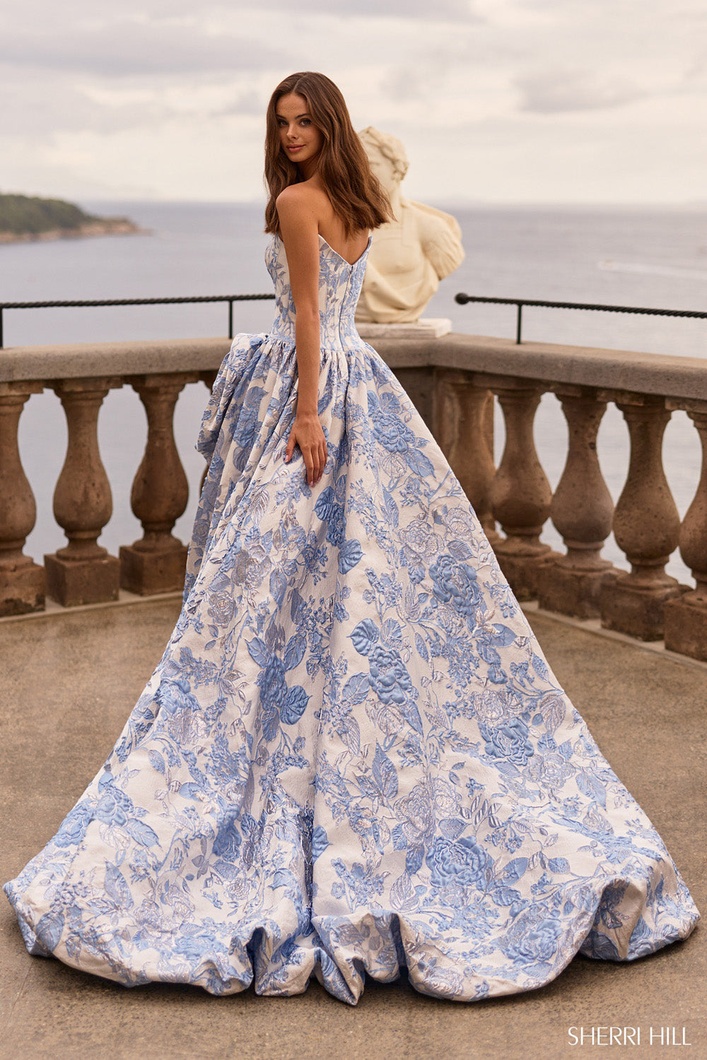 Sherri Hill 58092 strapless blue-and-white brocade ball gown, side pose leaning on stone railing near classical statue