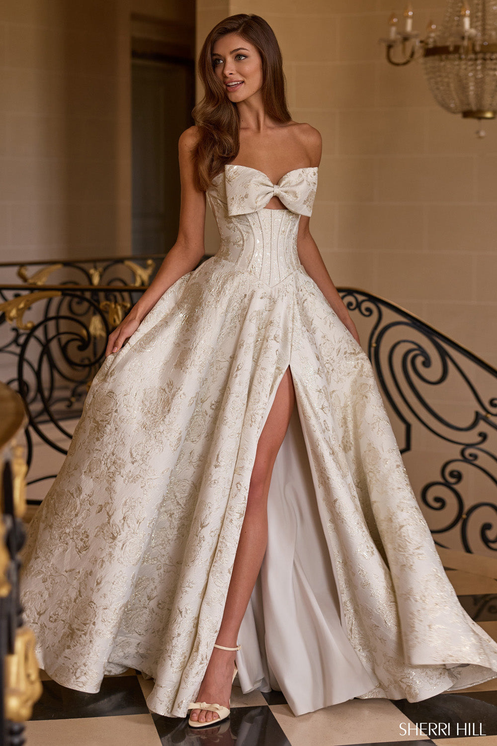 Sherri Hill 58240 in sand brocade ballgown with bow bust, corset bodice, and high-slit skirt, front view on grand staircase.