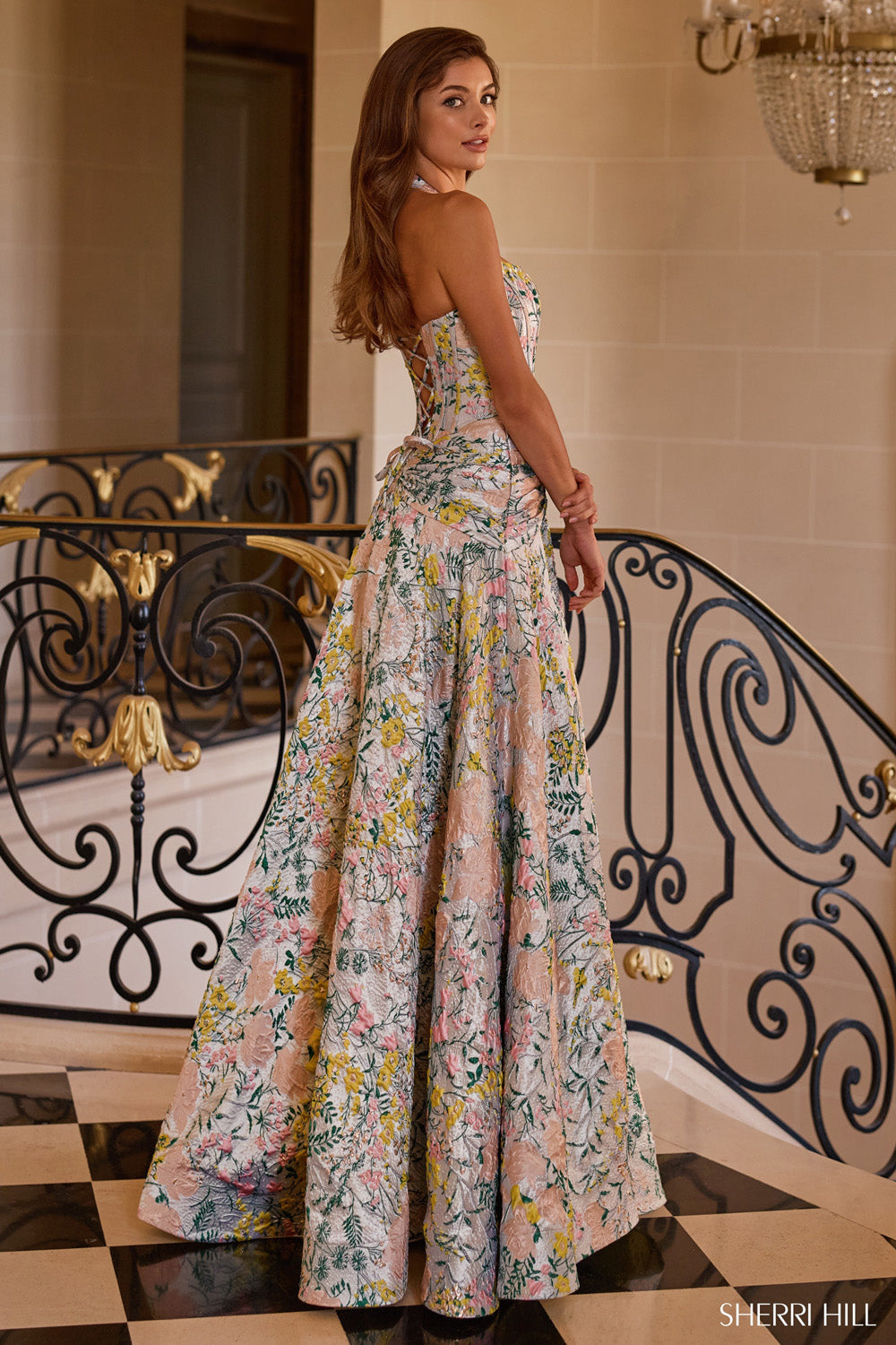 Back view of Sherri Hill 58284 showing lace-up open corset back and sweeping A-line floral brocade skirt on staircase.